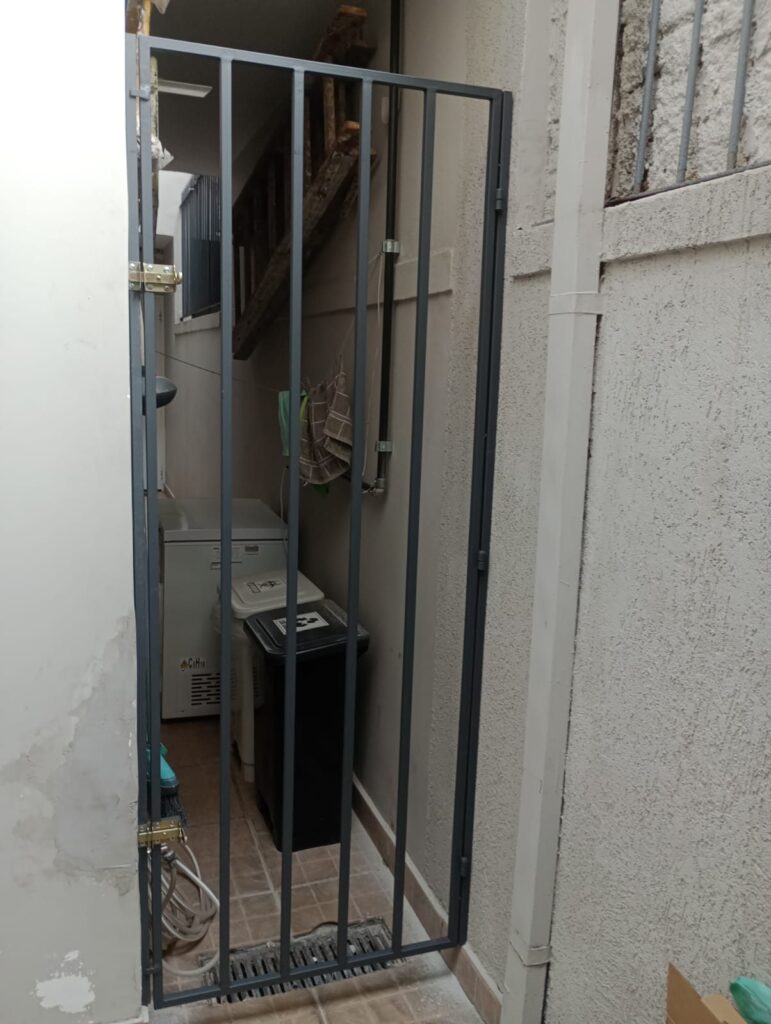 Portão social gradeado cinza instalado em corredor de residência em Moema, São Paulo